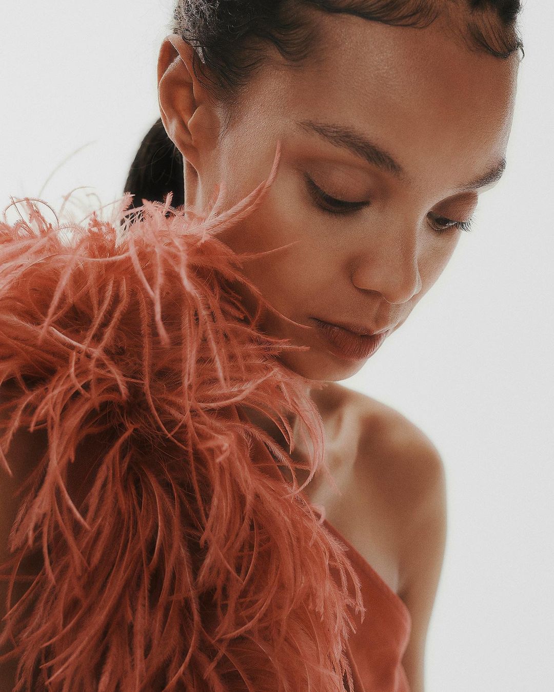 Close-up portrait of a woman with dark hair and feathered shoulder detail, looking down against a soft white backdrop in a beauty shot.