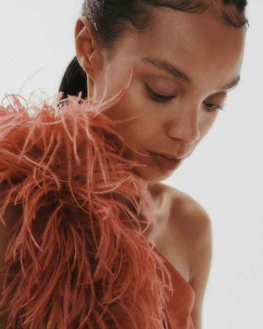 Close-up portrait of a woman with dark hair and feathered shoulder detail, looking down against a soft white backdrop in a beauty shot.