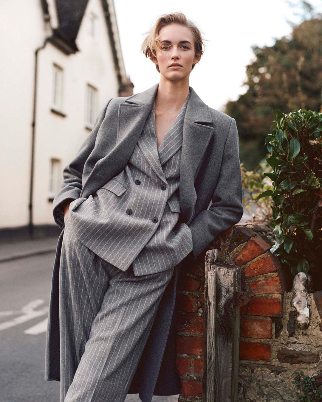 A woman in a grey pinstripe suit and long coat poses by a brick fence outdoors, with a white house visible in the background.