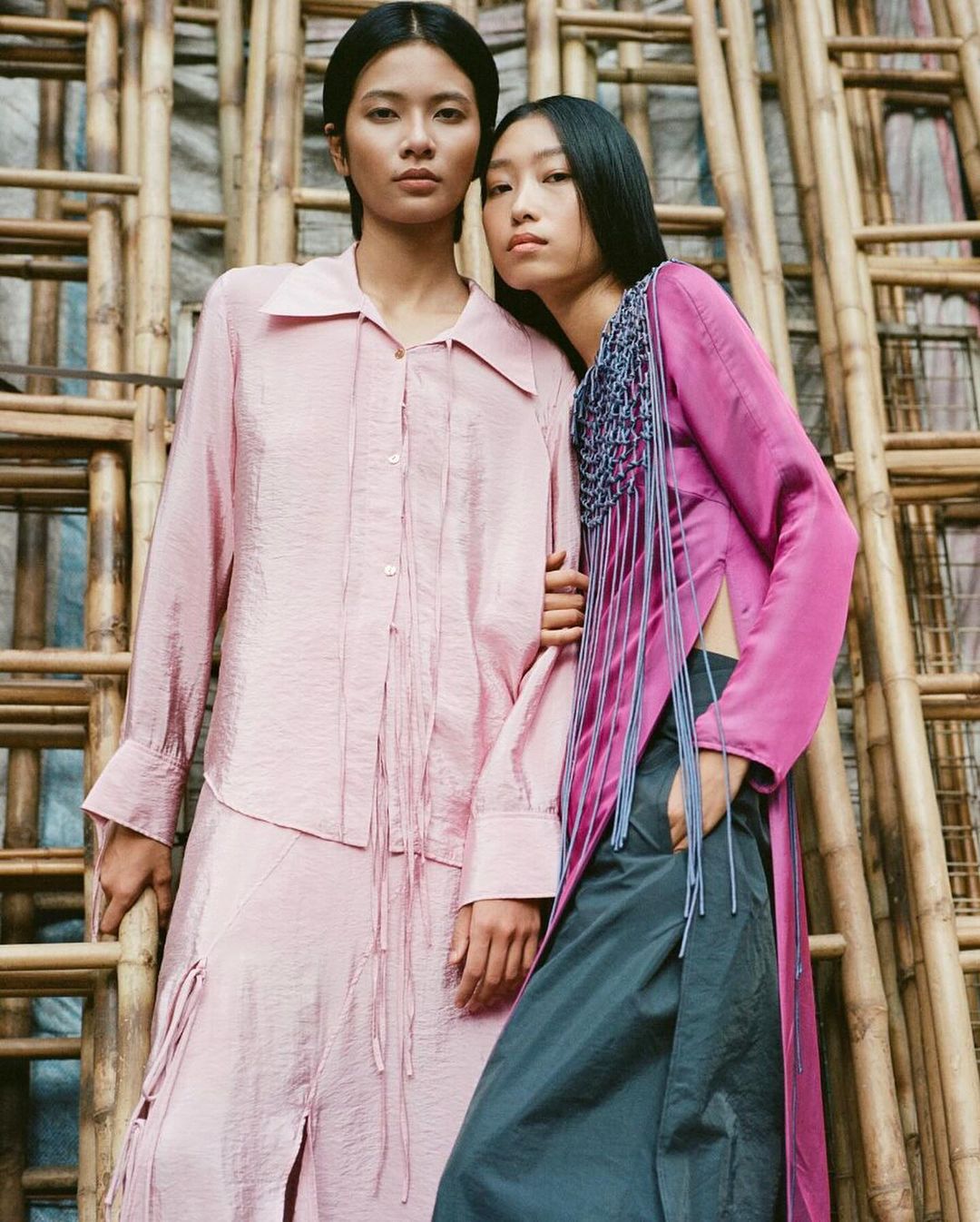 Two Asian female fashion models in pink and grey silk outfits pose indoors against a bamboo ladder structure.