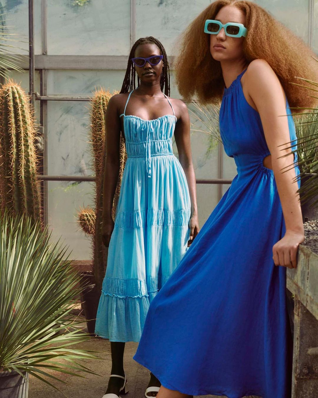Two women in colorful dresses pose in a greenhouse with tropical plants. One wears blue, the other light blue, both with sunglasses.