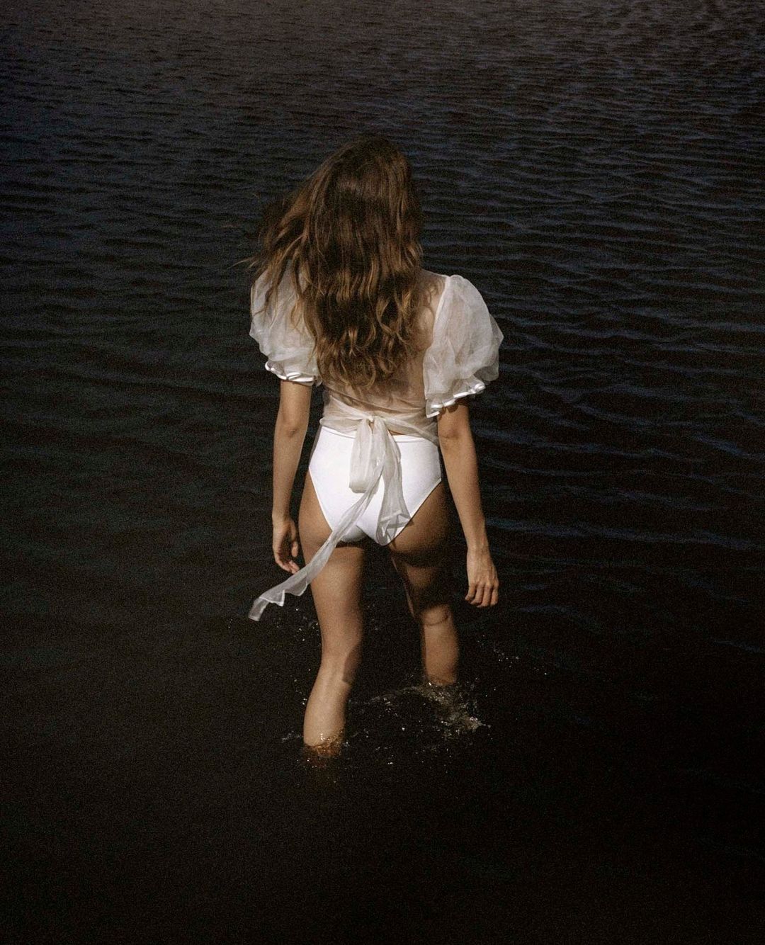A woman in a white swimsuit and sheer top stands in dark water, with her back to the camera, creating a serene and stylish image.