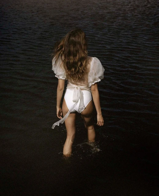 A woman in a white swimsuit and sheer top stands in dark water, with her back to the camera, creating a serene and stylish image.