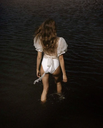 A woman in a white swimsuit and sheer top stands in dark water, with her back to the camera, creating a serene and stylish image.