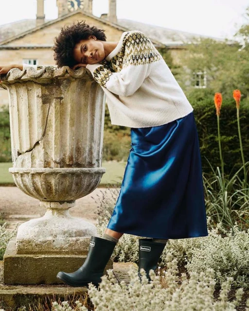 An adult woman with curly hair wearing a patterned sweater, satin skirt, and rain boots poses outdoors in front of a stone building.