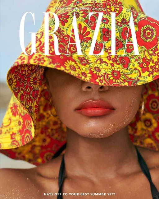 Close-up shot of a woman with a red and yellow floral hat that covers her eyes, red lipstick, and a black bikini top, set on a beach.
