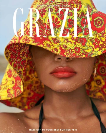 Close-up shot of a woman with a red and yellow floral hat that covers her eyes, red lipstick, and a black bikini top, set on a beach.