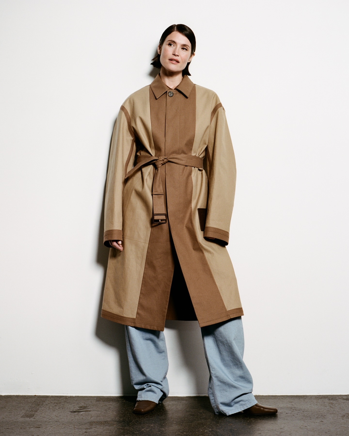 Model wearing a two-tone tan and brown belted coat with light blue trousers against a plain white wall