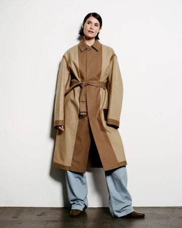 Model wearing a two-tone tan and brown belted coat with light blue trousers against a plain white wall