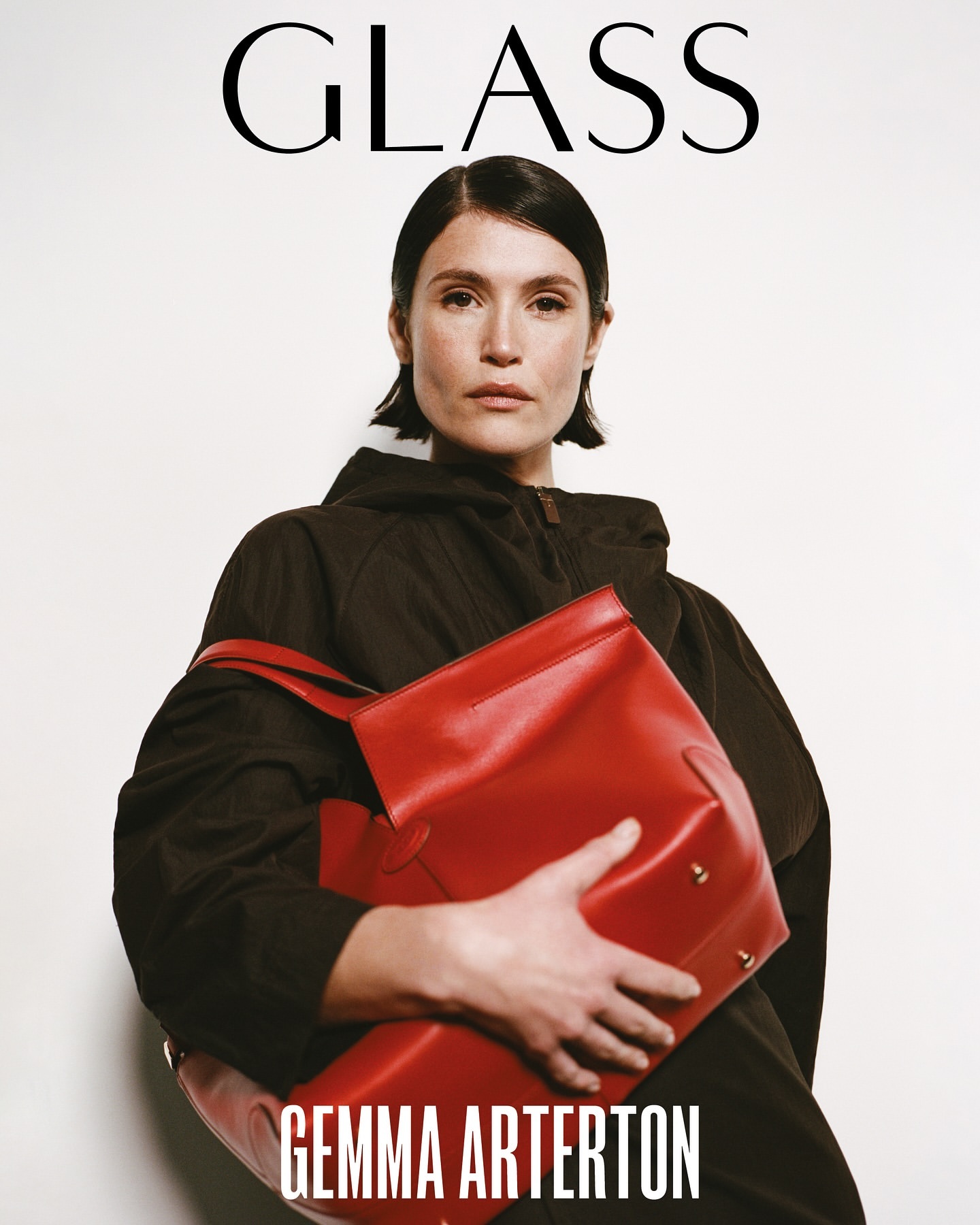 Gemma Arterton poses with a large red leather tote bag against a white background wearing a dark hooded jacket and neutral makeup.