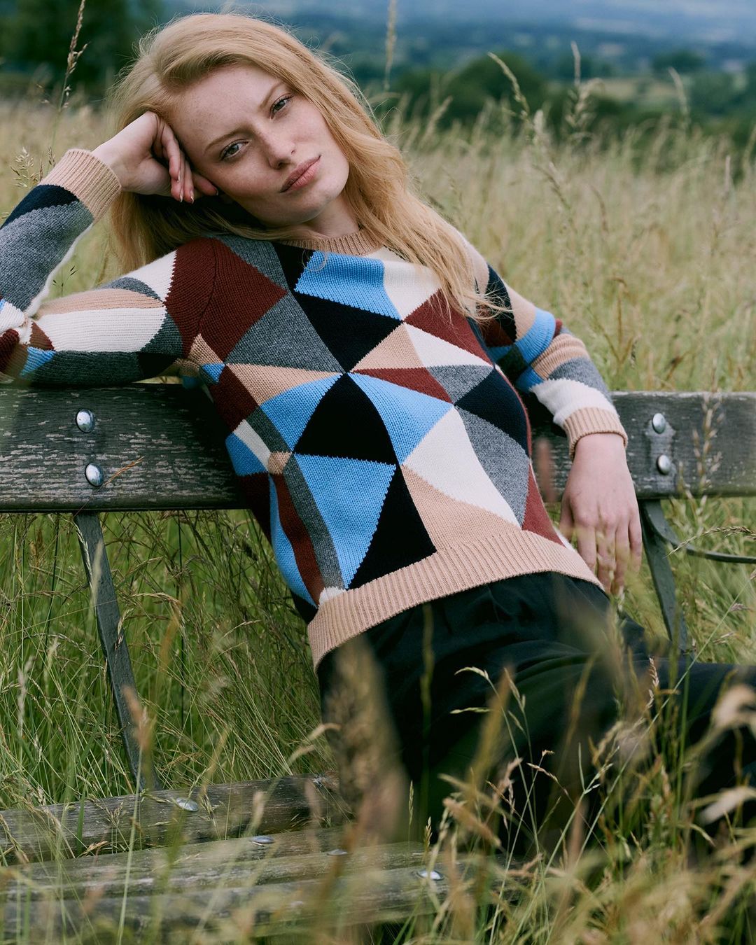 Fair-haired woman wearing geometric patterned knit sweater seated on outdoor wooden bench in tall meadow grass