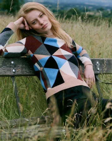 Fair-haired woman wearing geometric patterned knit sweater seated on outdoor wooden bench in tall meadow grass