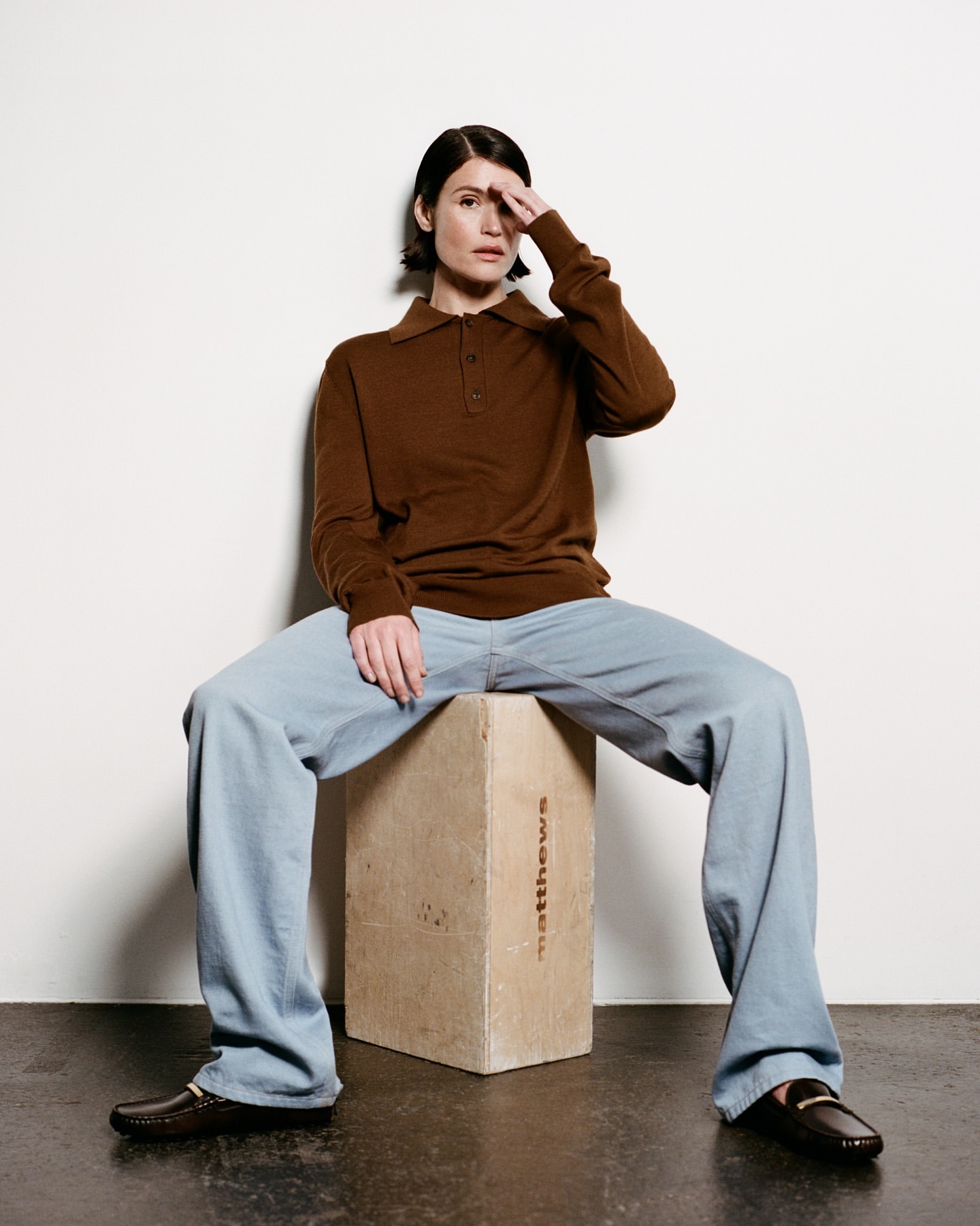 Adult woman in a brown knit top and light blue pants sits on a concrete block in a studio setting, striking a casual pose.