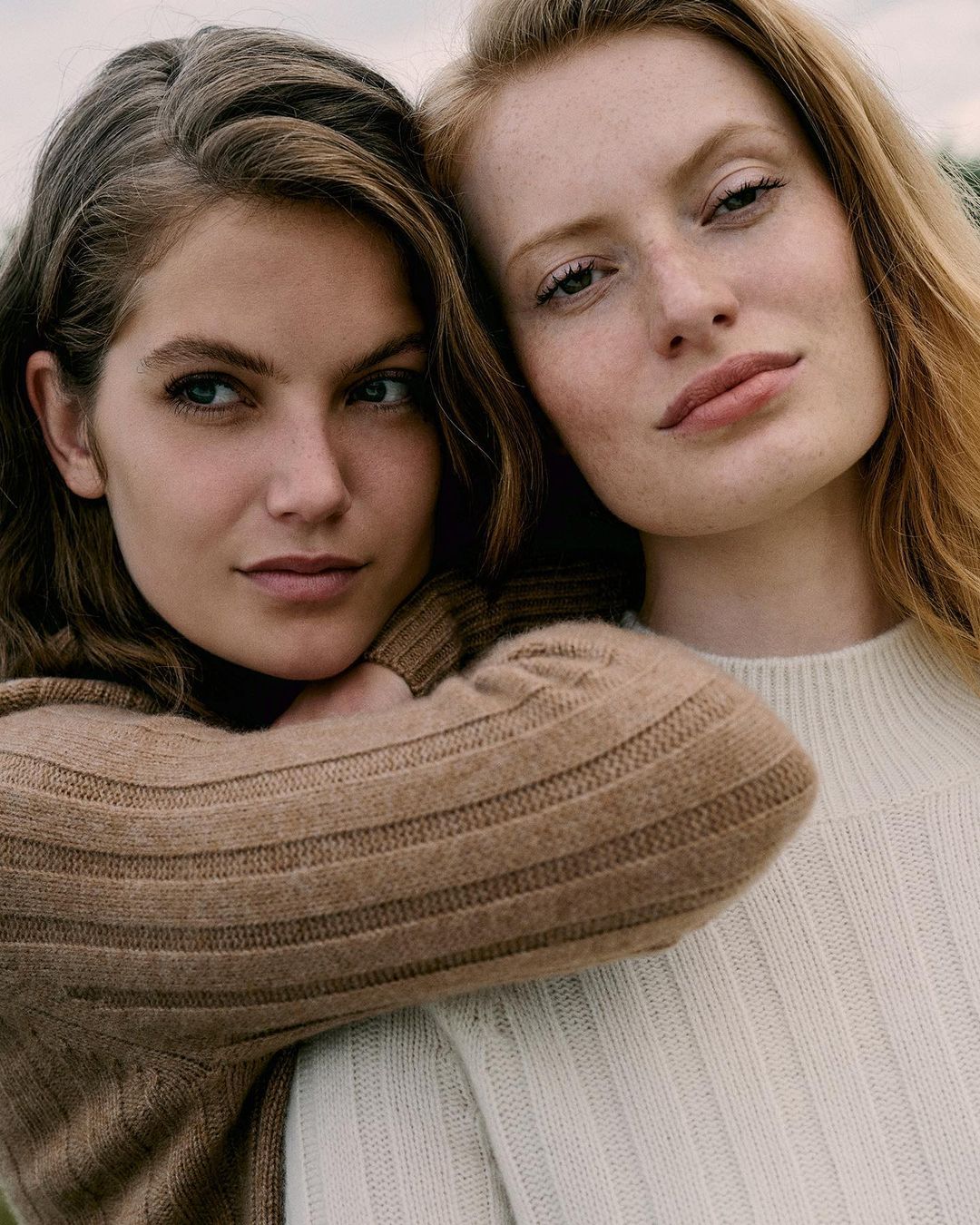 Portrait of two fair-skinned young women in close-up, both with neutral expressions and wearing tan and off-white sweaters, outdoor setting.