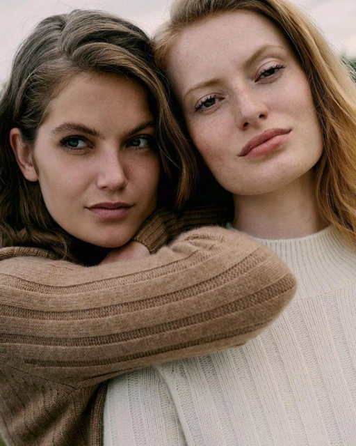 Portrait of two fair-skinned young women in close-up, both with neutral expressions and wearing tan and off-white sweaters, outdoor setting.