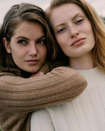 Portrait of two fair-skinned young women in close-up, both with neutral expressions and wearing tan and off-white sweaters, outdoor setting.
