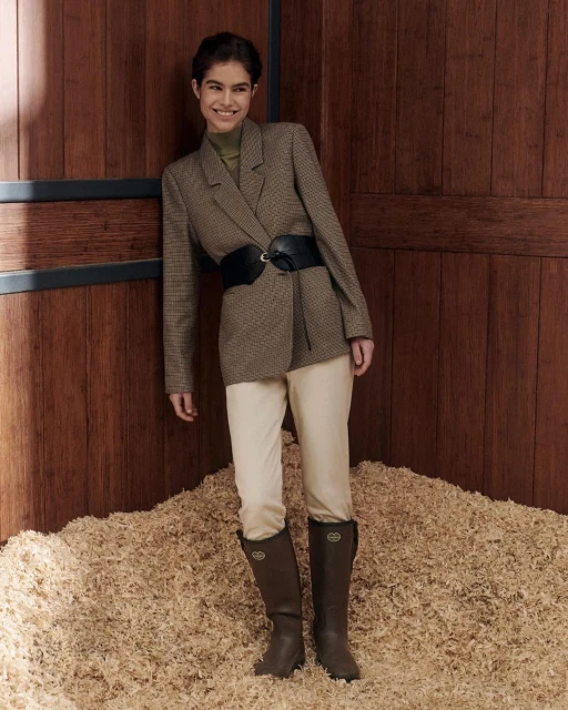 A woman in a blazer, white pants, and brown boots poses in front of a wooden wall and a pile of wood shavings.
