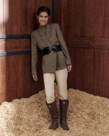 A woman in a blazer, white pants, and brown boots poses in front of a wooden wall and a pile of wood shavings.