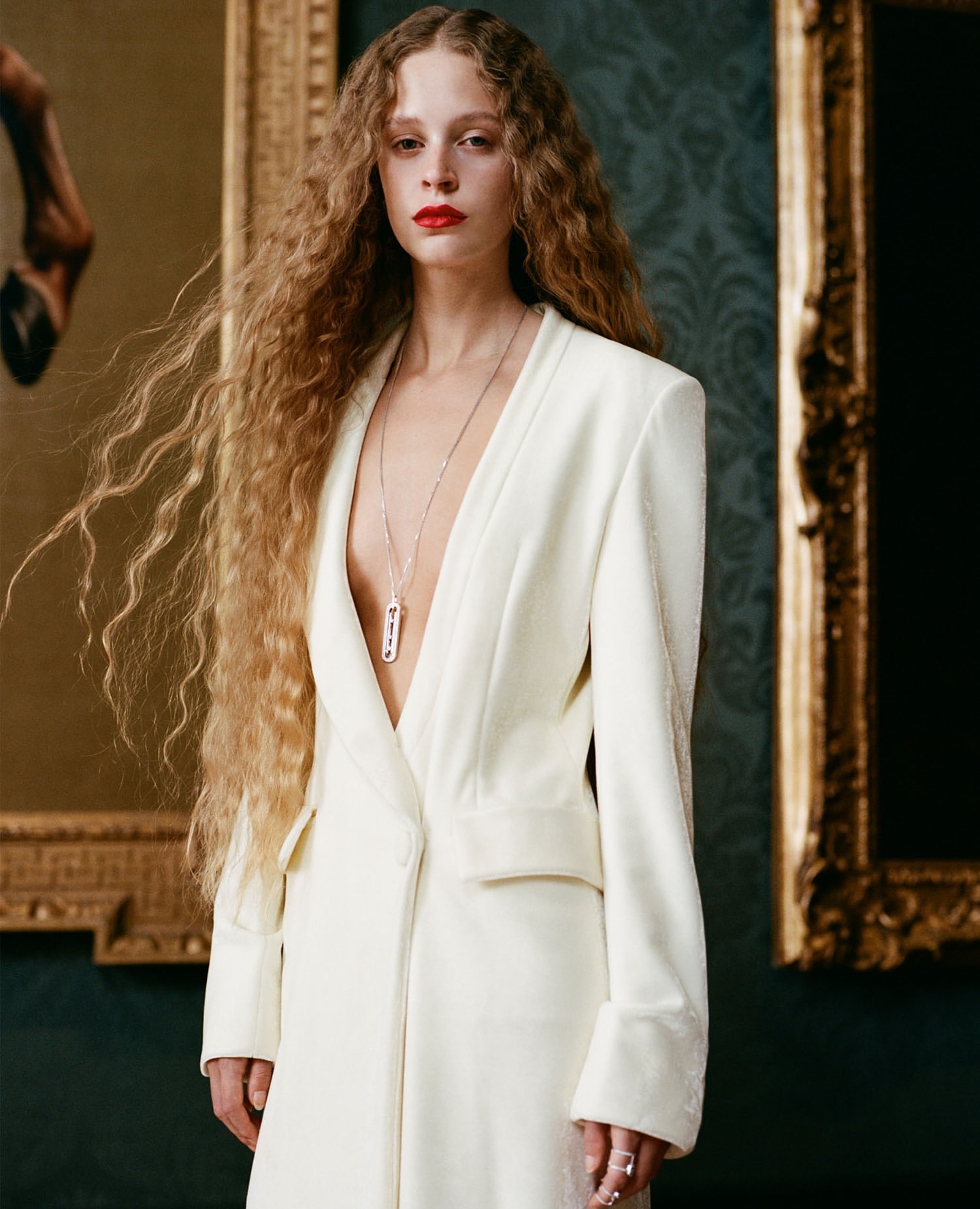 Woman with long curly blonde hair wearing a white blazer and pendant necklace against a dark patterned wall with framed art.
