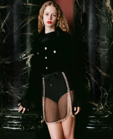 Woman with curly blonde hair poses in a black velvet blazer with a decorative skirt and column in the background. Fashion photography.