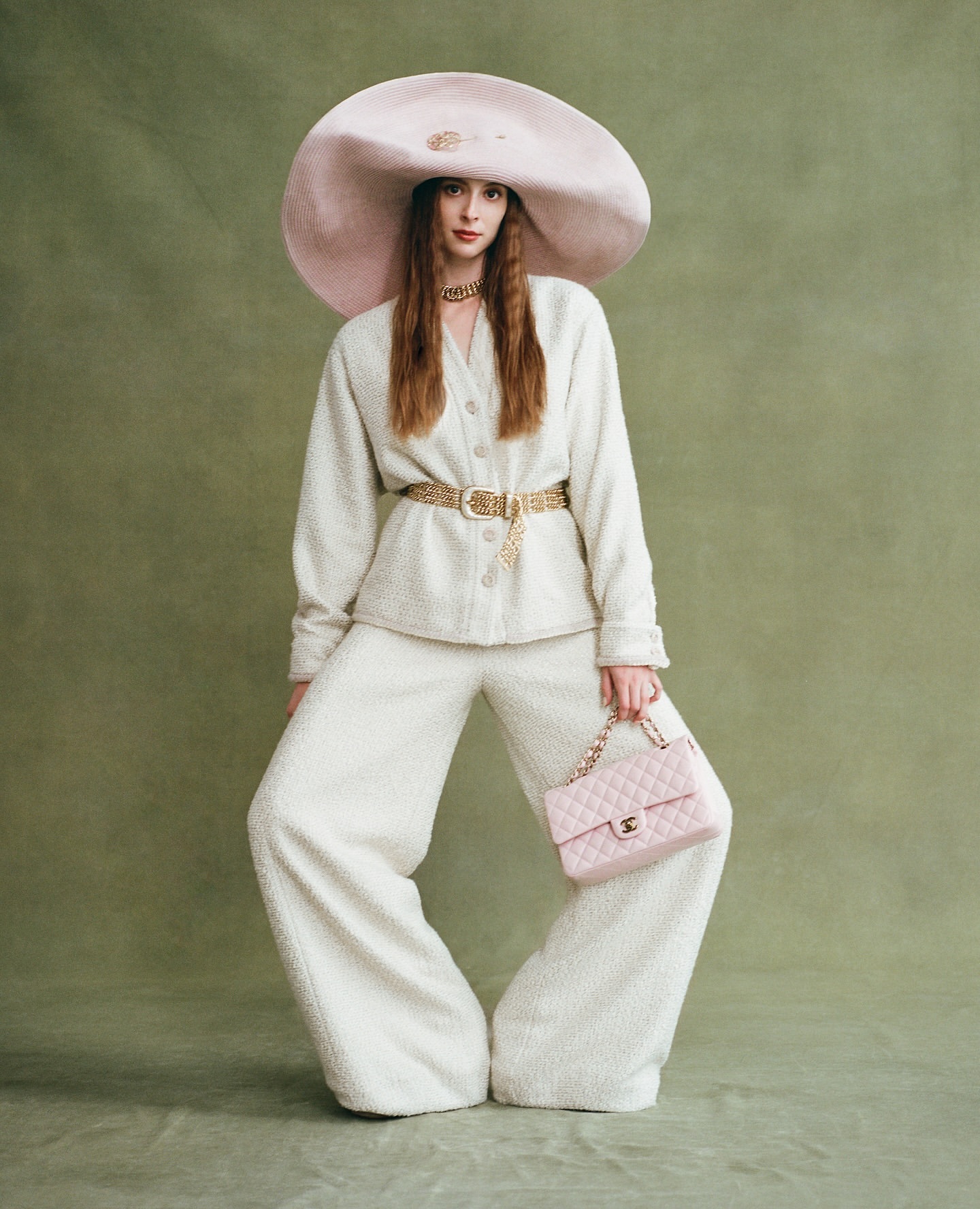 Fashionable woman poses in white suit and oversized hat holding pink purse against an olive green backdrop, style, studio fashion portrait.