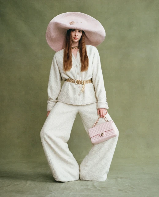 Fashionable woman poses in white suit and oversized hat holding pink purse against an olive green backdrop, style, studio fashion portrait.