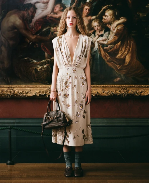 A woman with curly hair poses in a dress with a purse in front of an old painting in a gallery, creating a vintage fashion photograph.