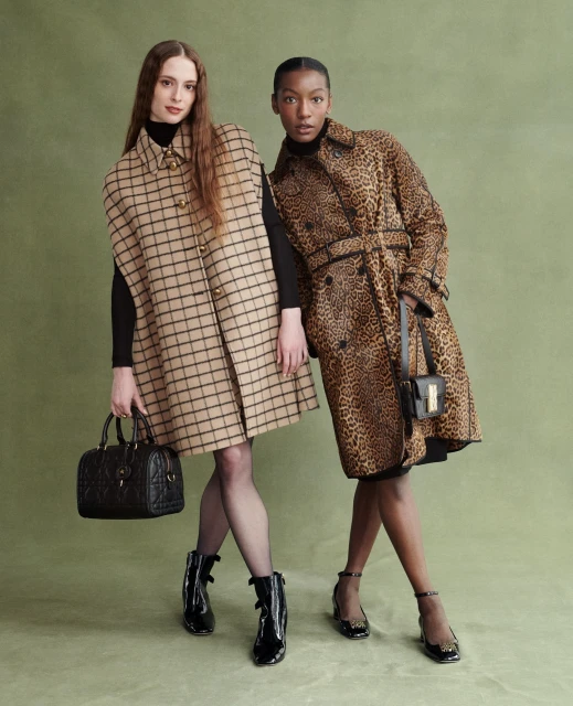 Two fashion models in coats, one in tan plaid and the other in leopard print, stand against a green backdrop, accessorized with purses and ankle boots.