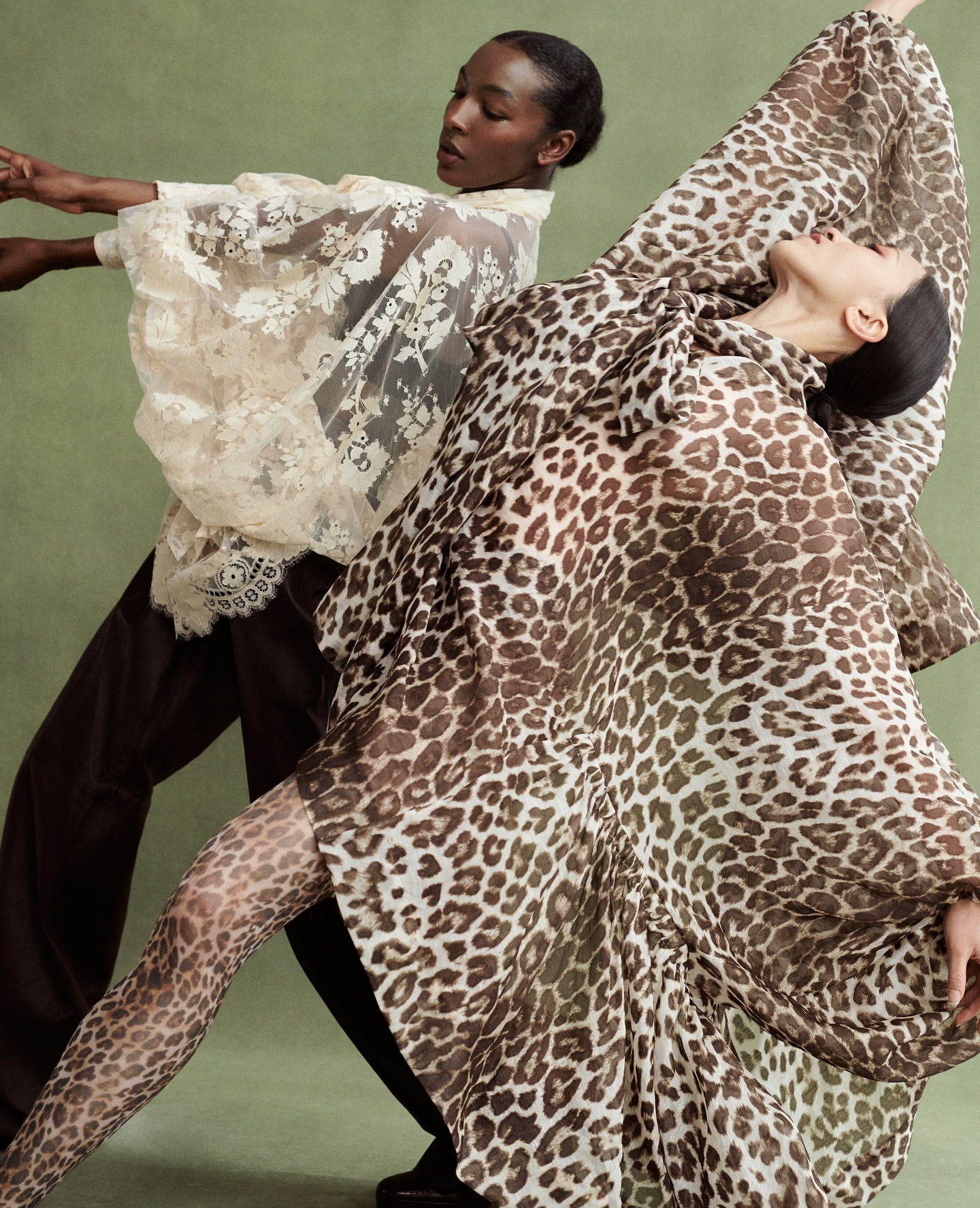 Two women pose wearing sheer animal print and lace tops against a green backdrop in a fashion editorial style, full studio lighting.