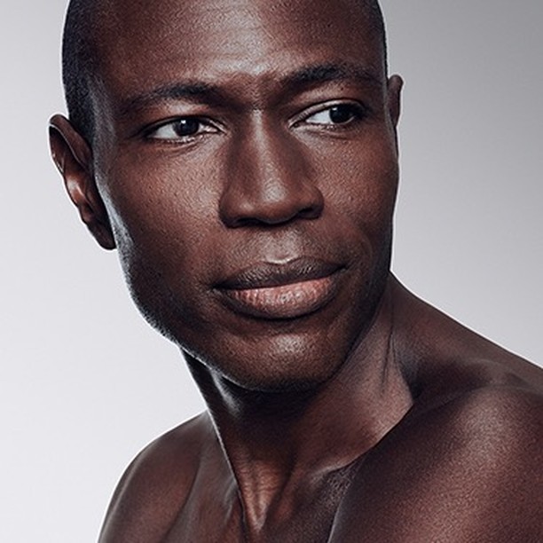 Close-up portrait of a handsome dark-skinned adult male looking off to the side against a gradient white to gray background, studio lit.