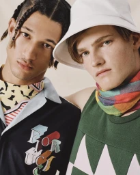 Two male models posing closely together wearing colourful patterned tops and a white bucket hat against a neutral background