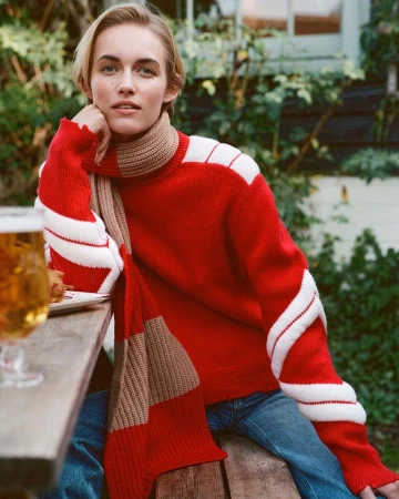 A blond woman wearing a red turtleneck sweater with white stripes and a matching scarf, seated outdoors at a wooden table.