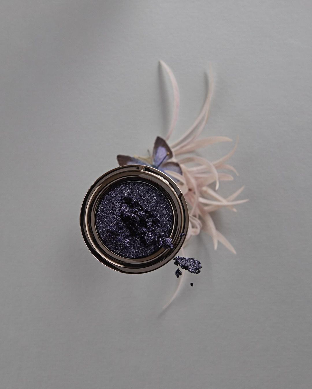 Overhead studio shot of purple eye shadow in silver container with decorative feathers on grey background for beauty advertisement.