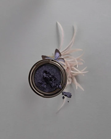 Overhead studio shot of purple eye shadow in silver container with decorative feathers on grey background for beauty advertisement.