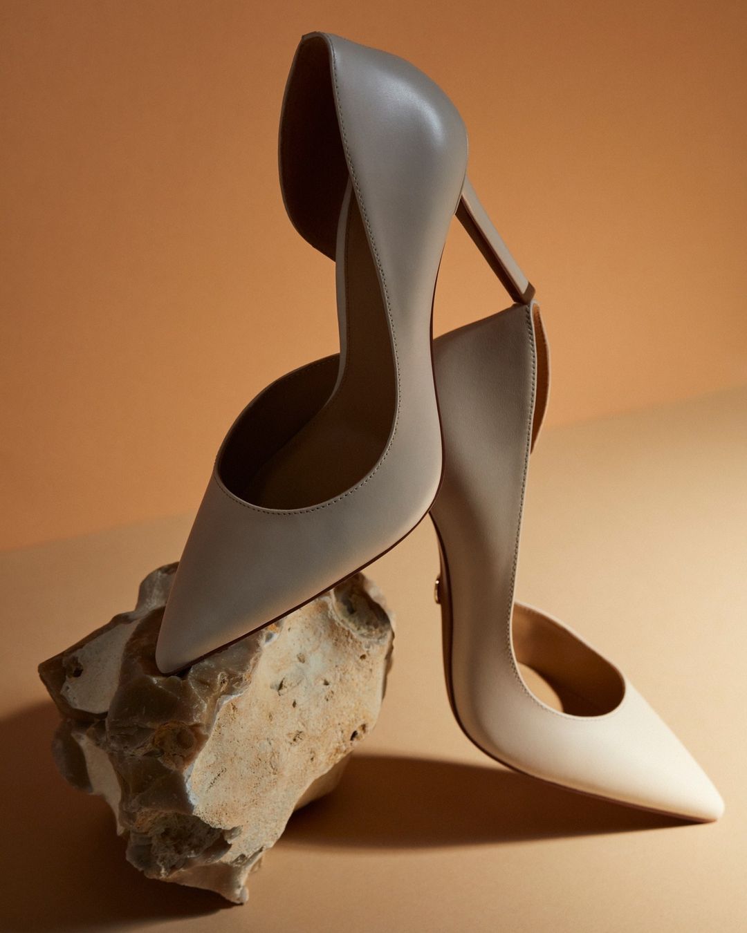 Elegant cream-colored high heel shoes are displayed atop a textured rock against a soft, monochromatic beige backdrop. Fashion still life.