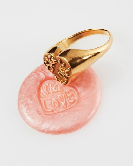 Close-up studio shot of a gold signet ring on a pink, heart-shaped wax seal with 'Big Love' text, against a white background.