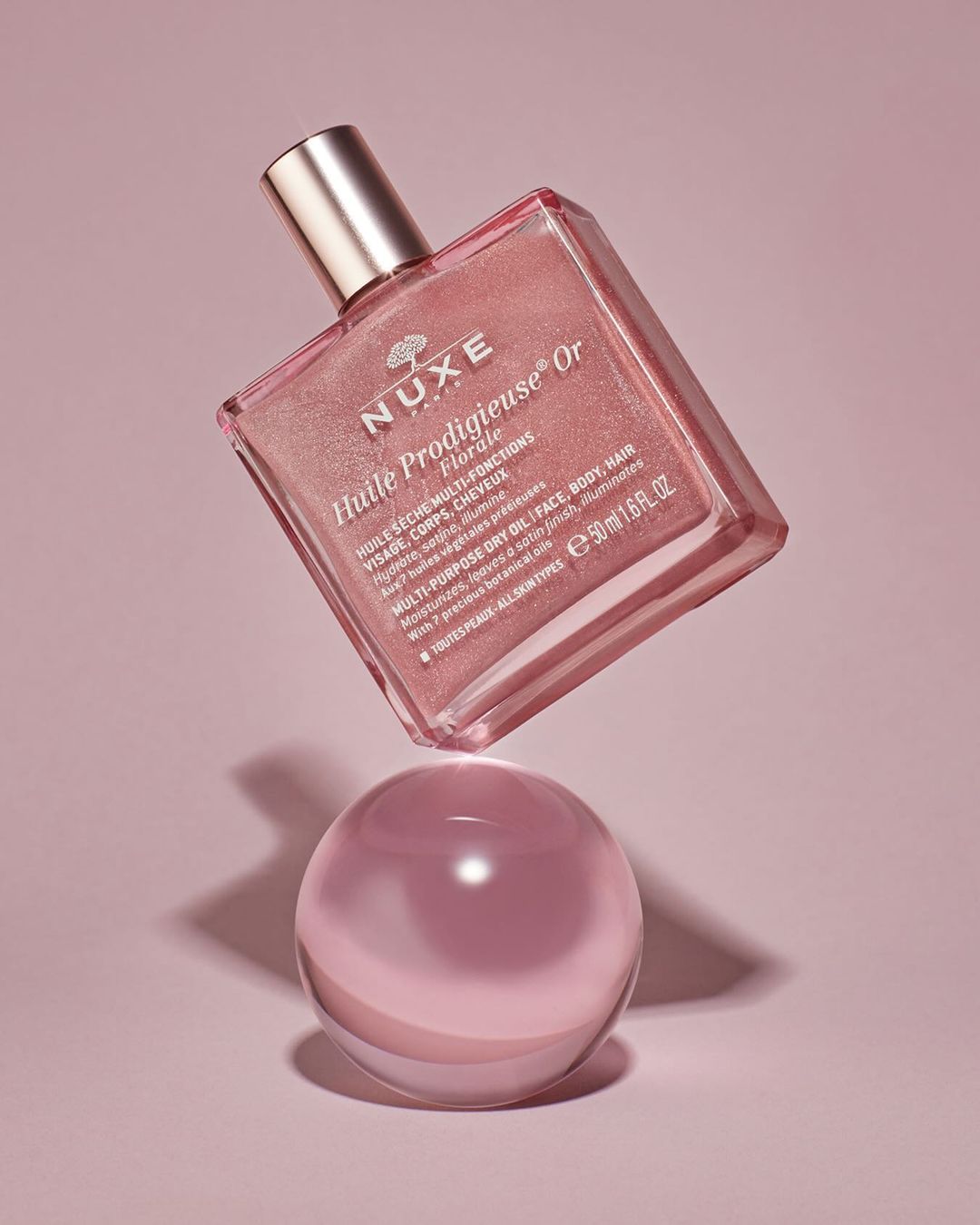 A pink and gold bottle of Nuxe Huile Prodigieuse Or Florale beauty oil floats above a clear sphere against a pink backdrop.