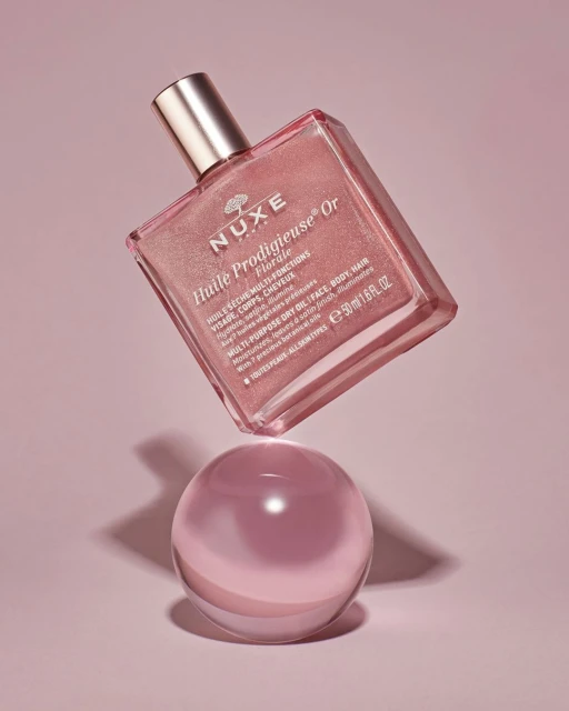 A pink and gold bottle of Nuxe Huile Prodigieuse Or Florale beauty oil floats above a clear sphere against a pink backdrop.