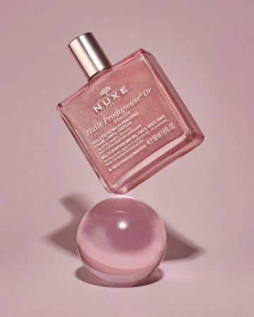 A pink and gold bottle of Nuxe Huile Prodigieuse Or Florale beauty oil floats above a clear sphere against a pink backdrop.
