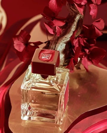 Still life of a clear glass perfume bottle with a red cap, set against a red and light-brown background with floral and branch accents.