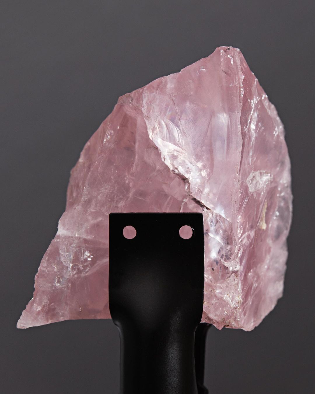 Close-up studio shot of a translucent rose quartz crystal on a black stand against a grey background, showcasing its texture and color.