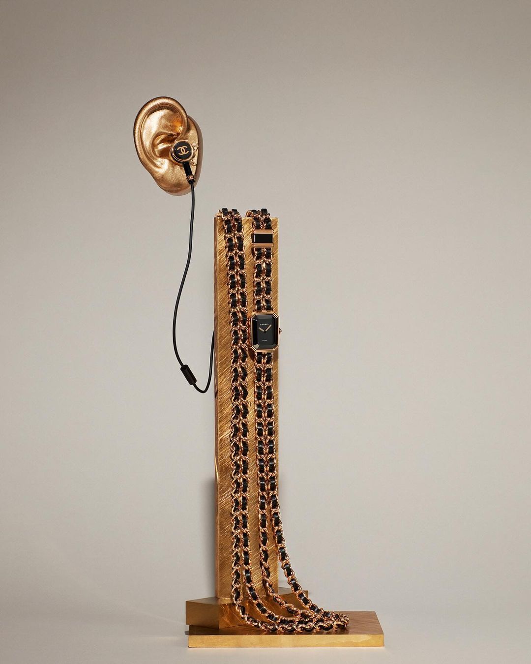 Conceptual shot of a gold ear sculpture with headphones, gold column with a black dial watch on a gold chain.