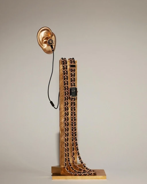 Conceptual shot of a gold ear sculpture with headphones, gold column with a black dial watch on a gold chain. 