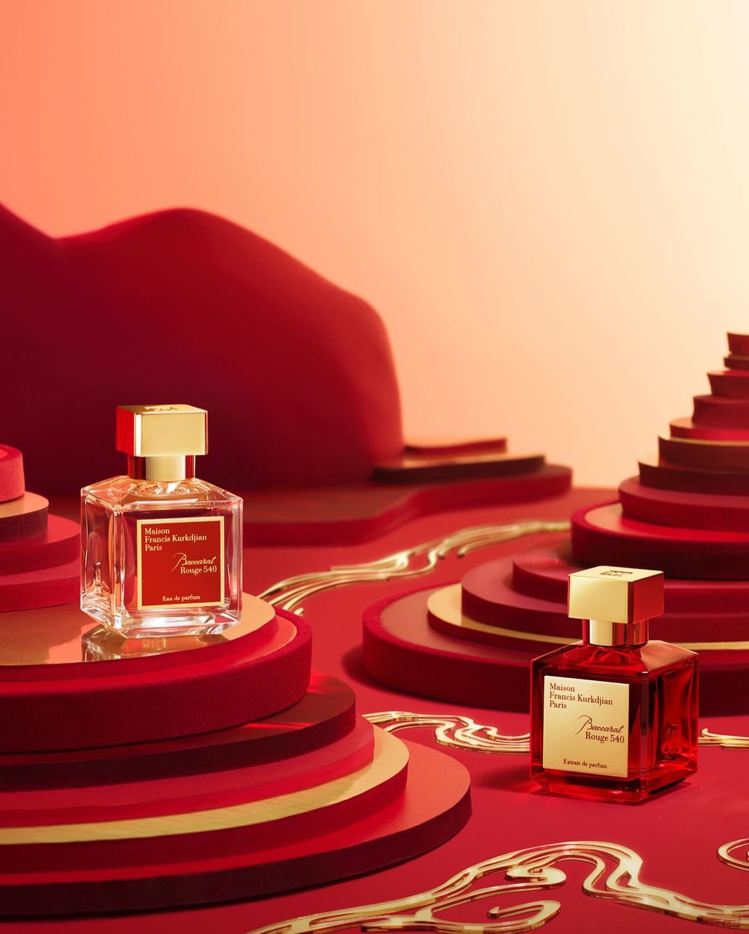 Two Baccarat Rouge 540 perfume bottles with gold accents sit on layered red platforms against a graduated light-orange background.