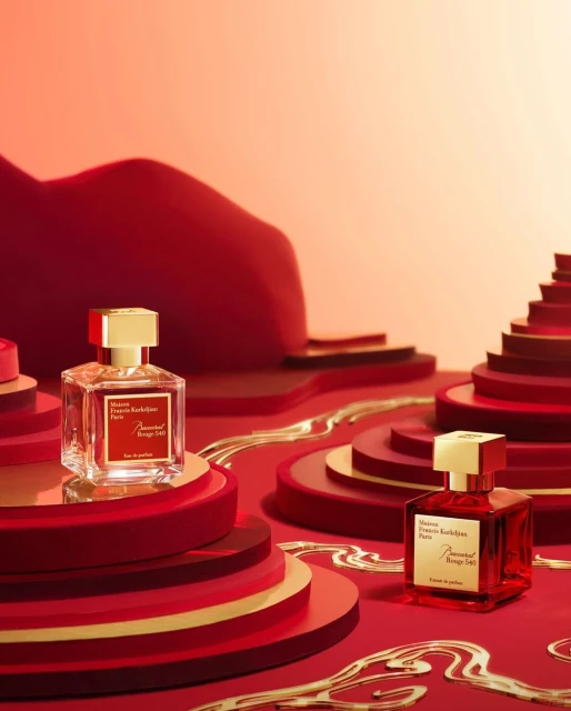 Two Baccarat Rouge 540 perfume bottles with gold accents sit on layered red platforms against a graduated light-orange background.