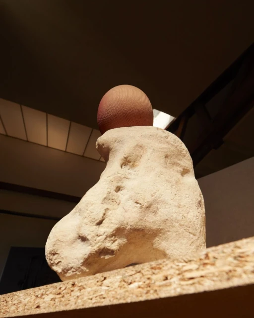 Abstract sculpture of a rounded light-colored stone base with a peach-colored mesh sphere on top, displayed indoors on a wood surface.