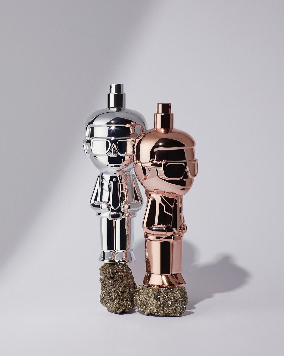 Two shiny, stylized figurine perfume bottles, one silver and one copper, sit on metallic pyrite rocks against a gradient white background.