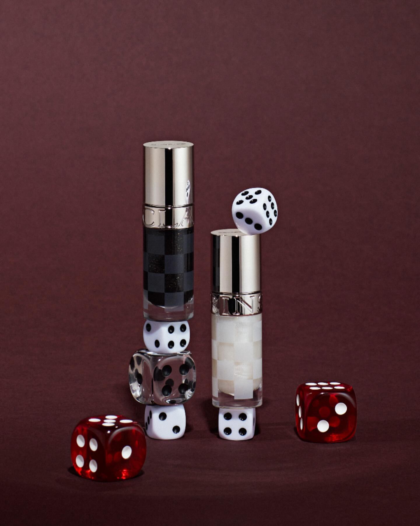 Two beauty product bottles surrounded by dice on a maroon background create an arrangement for cosmetic promotion.
