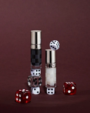 Two beauty product bottles surrounded by dice on a maroon background create an arrangement for cosmetic promotion.