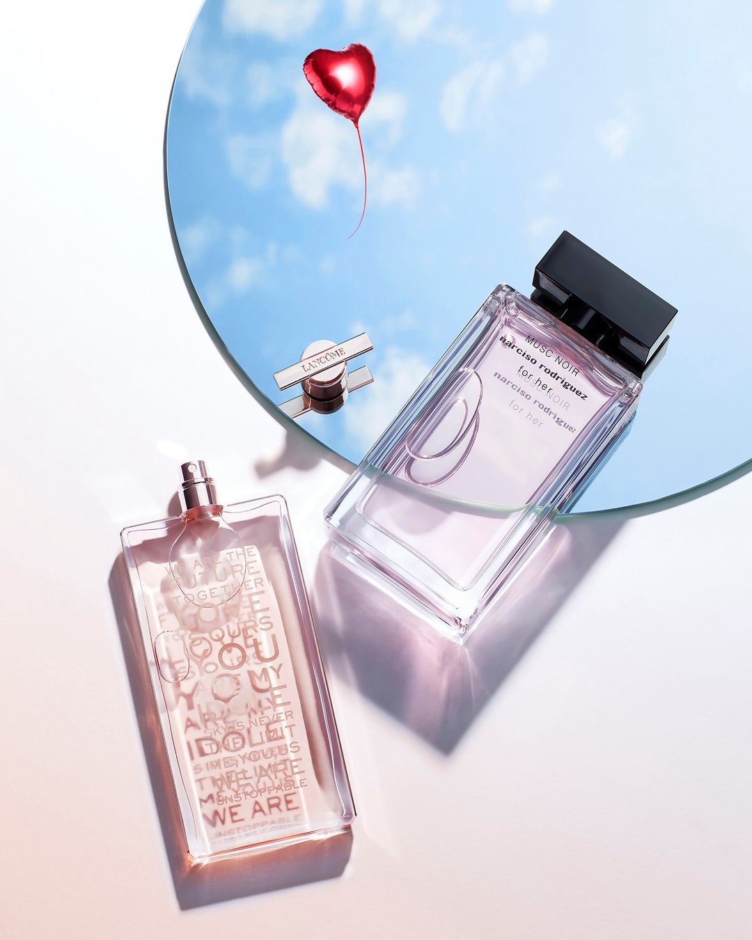 Two fragrance bottles, one with text inside, are displayed near a mirror reflecting a sky with a red heart-shaped balloon.